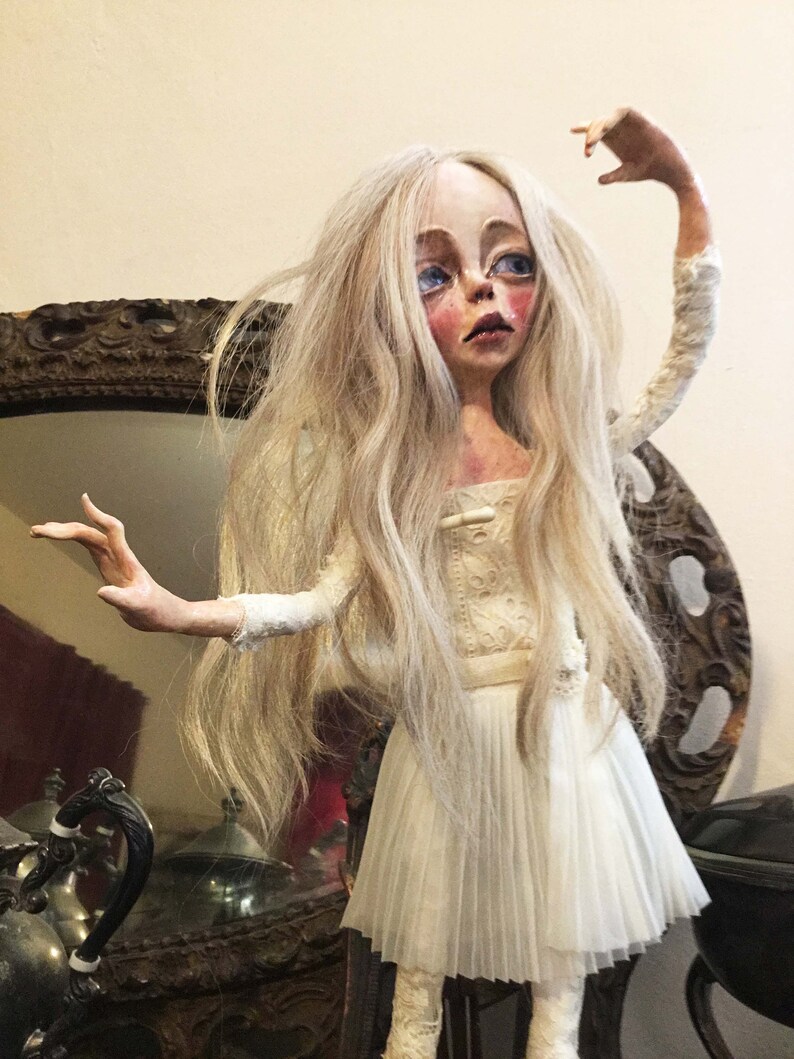 May include: A porcelain doll with long white hair, wearing a white dress with a lace bodice and a pleated skirt. The doll has a painted face with blue eyes and rosy cheeks.