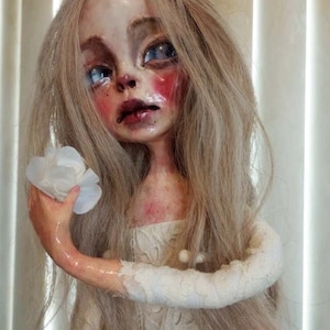 May include: A porcelain doll with long blonde hair, wearing a white lace dress and holding a white flower. The doll has a pale complexion, rosy cheeks, and blue eyes.