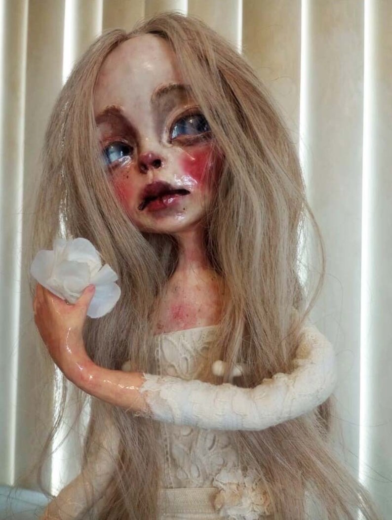 May include: A porcelain doll with long blonde hair, wearing a white lace dress and holding a white flower. The doll has a pale complexion, rosy cheeks, and blue eyes.