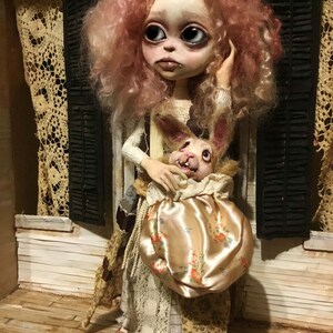 May include: A handcrafted doll with a pale face, large eyes, and pink, curly hair. The doll wears a patterned dress and holds a satin bag with a floral design, containing a rabbit figure. The doll stands on a wooden porch.