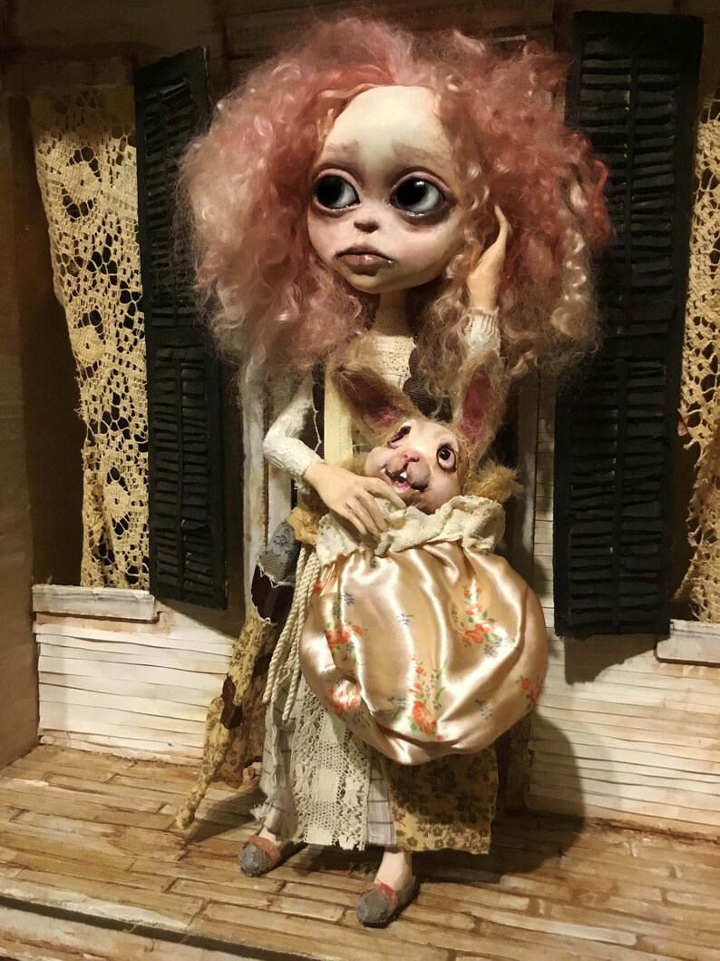 May include: A handcrafted doll with a pale face, large eyes, and pink, curly hair. The doll wears a patterned dress and holds a satin bag with a floral design, containing a rabbit figure. The doll stands on a wooden porch.