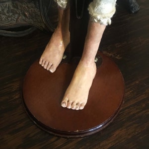 May include: Close-up of the feet of a doll with bare feet and a wooden base. The doll's feet are made of painted porcelain and have a realistic appearance.
