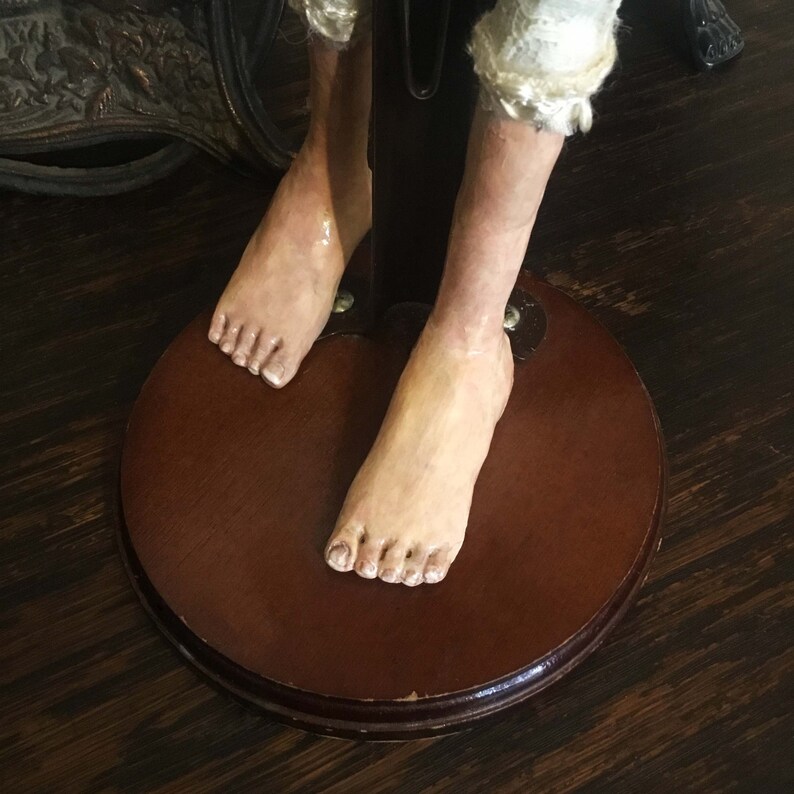 May include: Close-up of the feet of a doll with bare feet and a wooden base. The doll's feet are made of painted porcelain and have a realistic appearance.