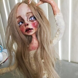 May include: A porcelain doll with long blonde hair, wearing a white lace dress. The doll has a pale complexion, blue eyes, and rosy cheeks.