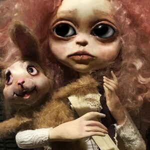 May include: A handmade doll with a pale face, large black eyes, and pink curly hair. The doll is holding a brown, furry rabbit. The doll's clothing includes a white lace sleeve and a brown ribbon.