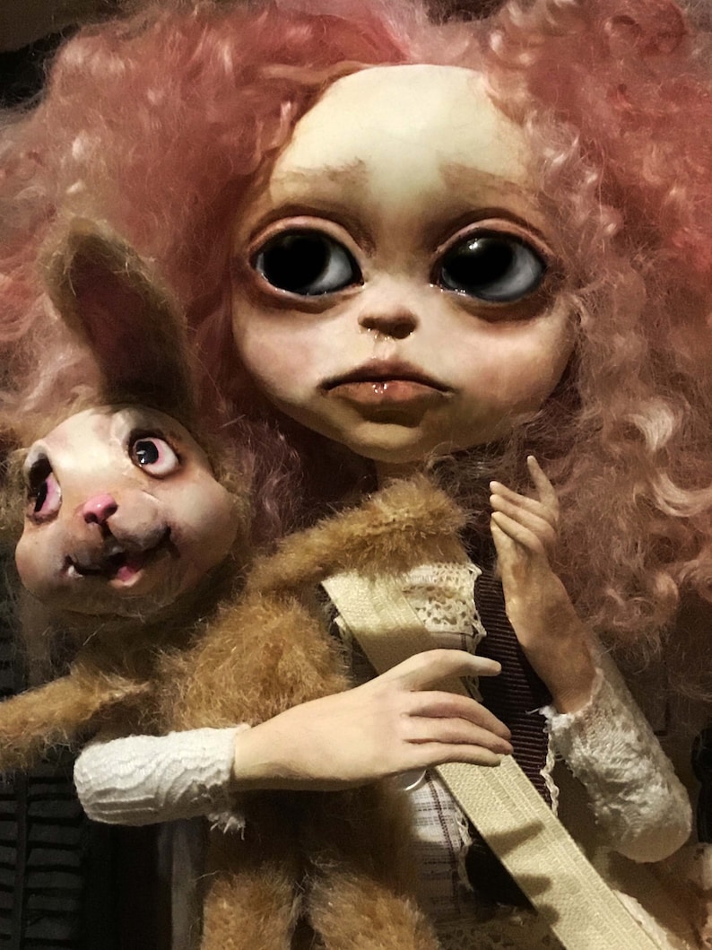 May include: A handmade doll with a pale face, large black eyes, and pink curly hair. The doll is holding a brown, furry rabbit. The doll's clothing includes a white lace sleeve and a brown ribbon.