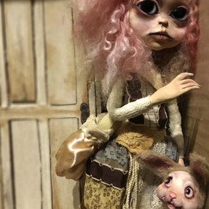 May include: A handmade doll with a pale face, large black eyes, and pink curly hair. The doll wears a patchwork outfit with a tan satchel and holds a small stuffed rabbit. The background features a white door.