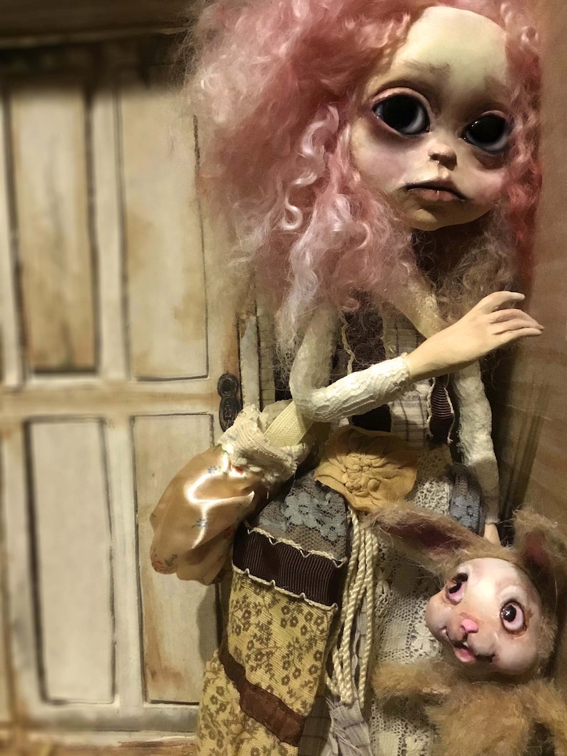 May include: A handmade doll with a pale face, large black eyes, and pink curly hair. The doll wears a patchwork outfit with a tan satchel and holds a small stuffed rabbit. The background features a white door.