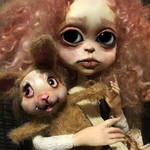 May include: A handmade doll with a pale face, large black eyes, and pinkish-red curly hair. The doll is holding a brown plush rabbit with pink accents. The doll is wearing a cream-colored outfit.