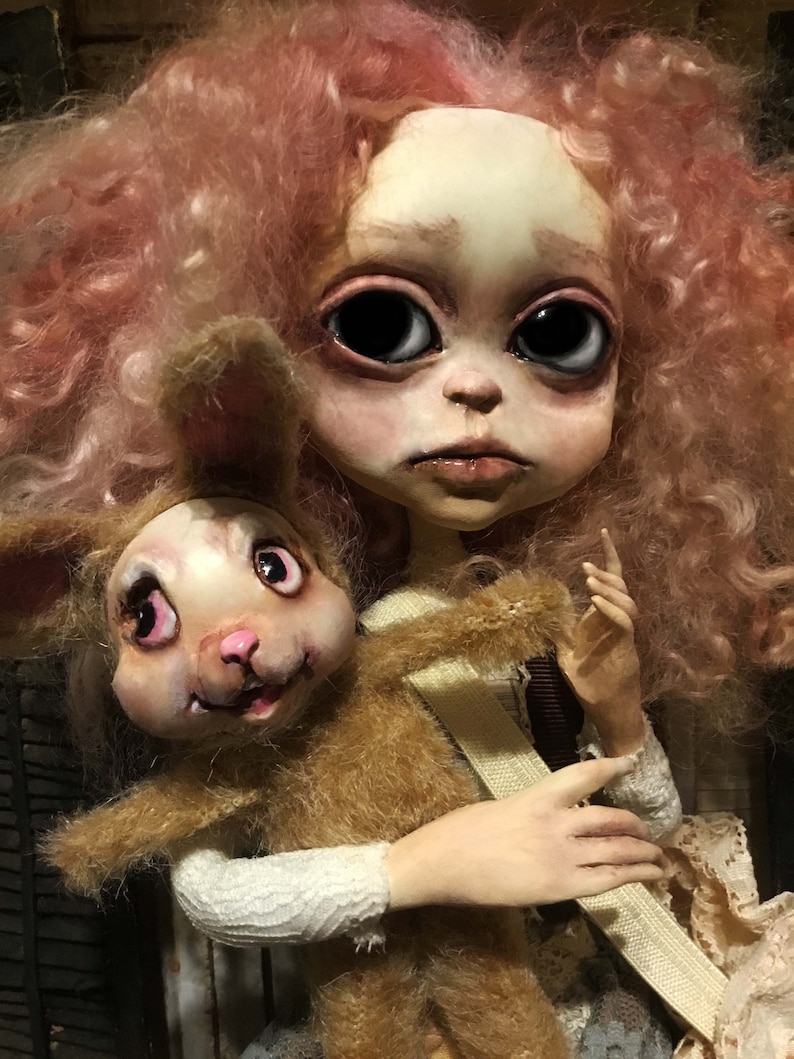 May include: A handmade doll with a pale face, large black eyes, and pinkish-red curly hair. The doll is holding a brown plush rabbit with pink accents. The doll is wearing a cream-colored outfit.