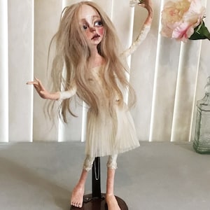 May include: A porcelain doll with long blonde hair, wearing a white dress and tights, holding a white flower. The doll is standing on a wooden base.