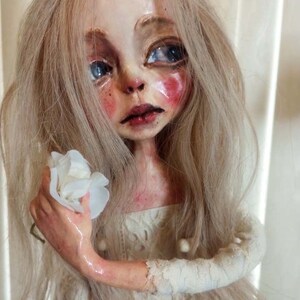 May include: A porcelain doll with long blonde hair, wearing a white lace dress and holding a white flower. The doll has a painted face with blue eyes and rosy cheeks.