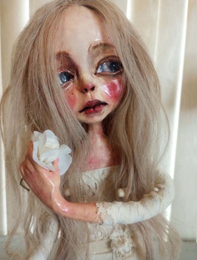 May include: A porcelain doll with long blonde hair, wearing a white lace dress and holding a white flower. The doll has a painted face with blue eyes and rosy cheeks.