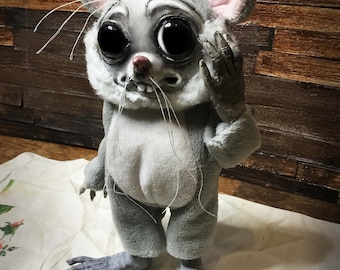 May include: A gray plush mouse doll with large black eyes and a toothy grin. The doll is wearing a gray hooded jumpsuit and has long, thin whiskers.
