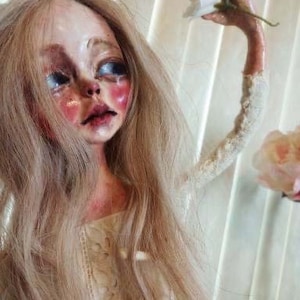 May include: A porcelain doll with long blonde hair, wearing a white lace dress. The doll has a pale complexion, rosy cheeks, and large blue eyes. One arm is outstretched, holding a white flower.