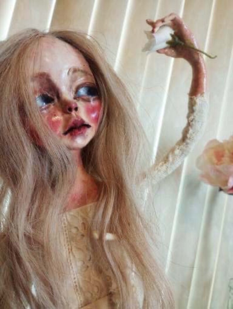 May include: A porcelain doll with long blonde hair, wearing a white lace dress. The doll has a pale complexion, rosy cheeks, and large blue eyes. One arm is outstretched, holding a white flower.