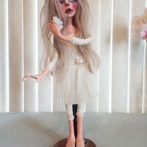 May include: A porcelain doll with long blonde hair, wearing a white dress and holding a white flower. The doll is standing on a wooden base.