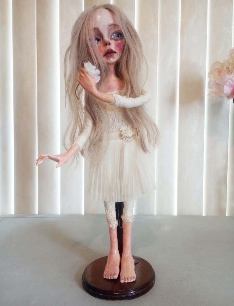May include: A porcelain doll with long blonde hair, wearing a white dress and holding a white flower. The doll is standing on a wooden base.