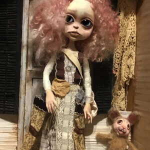 May include: A handmade doll with a pale face, large eyes, and pink curly hair. The doll wears a layered dress with lace and floral patterns, and a brown sash. A small stuffed rabbit with a creepy smile sits nearby.