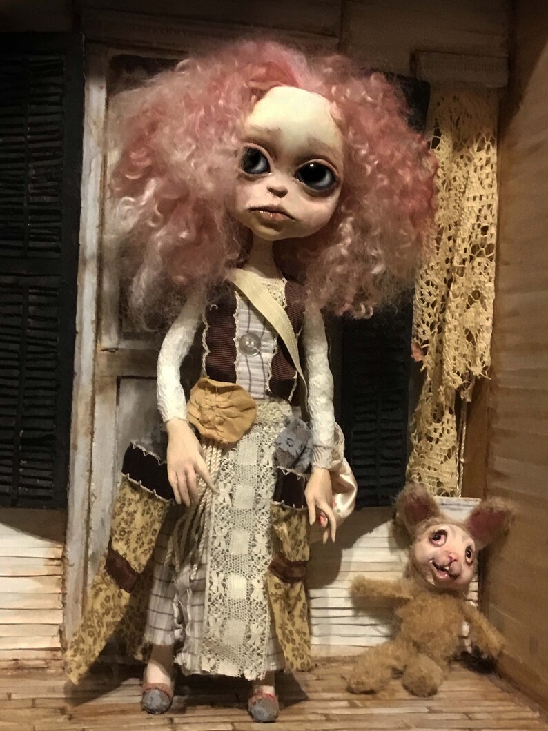 May include: A handmade doll with a pale face, large eyes, and pink curly hair. The doll wears a layered dress with lace and floral patterns, and a brown sash. A small stuffed rabbit with a creepy smile sits nearby.