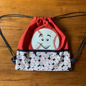 May include: Red drawstring bag with a white baseball face and a blue and white baseball pattern on the bottom.