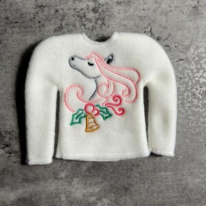 May include: A white long-sleeved sweater with a whimsical embroidered design. The design features a unicorn with pink hair, a bell, and holly. The sweater is made of a soft, fuzzy material, perfect for the holiday season.