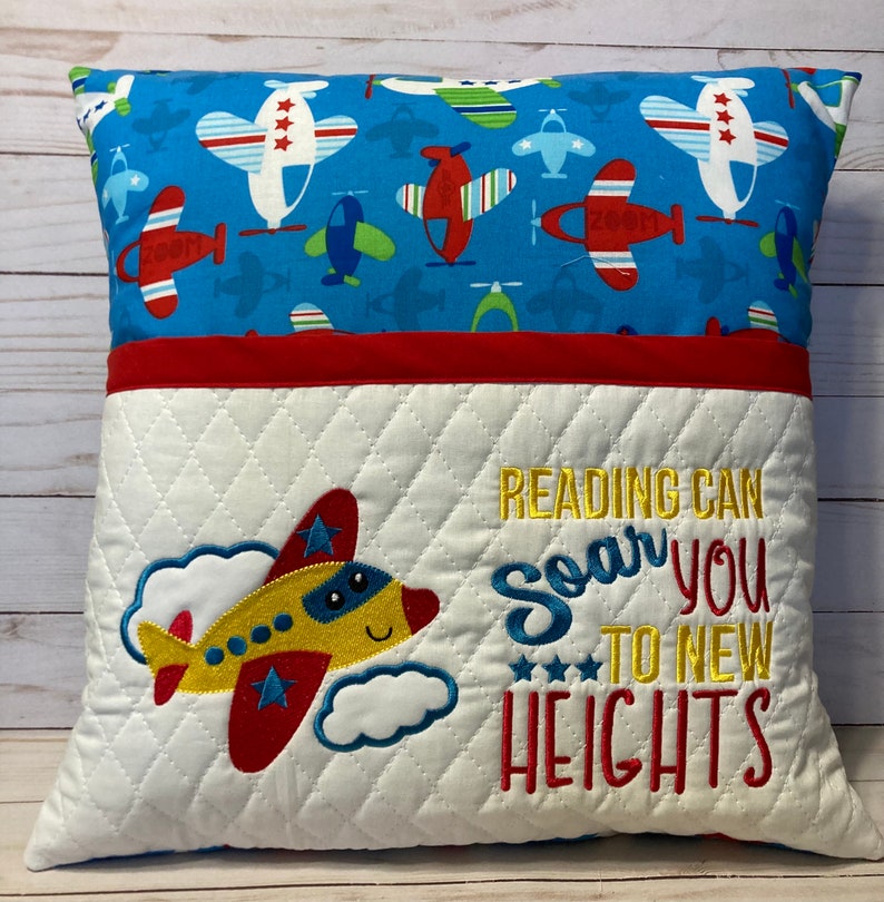 K&ouml;nnte beinhalten: Ein wei&szlig;es und rotes gestepptes Kissen mit einer Tasche auf der Vorderseite. Das Kissen hat einen blauen und wei&szlig;en Stoff auf der R&uuml;ckseite mit einem Muster aus Flugzeugen. Die Vorderseite des Kissens ist mit einem Flugzeug bestickt und dem Text "READING CAN SOAR YOU TO NEW HEIGHTS".