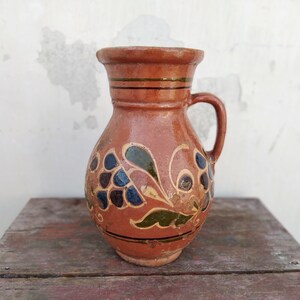 Op de afbeelding: Een bruine keramische kan met een handvat, versierd met een bloemmotief in tinten blauw, groen en goud.