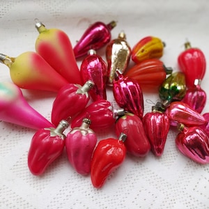 Puede incluir: Una colección de adornos navideños vintage en varias formas y colores. Los adornos incluyen gotas, pimientos y otros diseños frutales rojos, rosas y dorados. Están dispuestos sobre una superficie blanca texturizada.