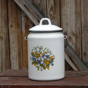 Könnte beinhalten: Weiße Emaille-Milchkanne mit einem Blumendesign mit blauen und gelben Blumen. Die Kanne hat einen schwarzen Deckel und einen Griff.