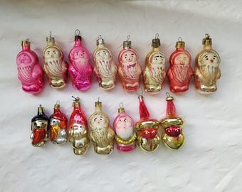 May include: A collection of vintage glass Christmas baubles featuring various characters, including Father Christmas, elves, and other festive figures. The baubles are mostly red, pink, and white with gold accents.