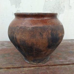 Puede incluir: Una olla de barro marrón con un esmalte marrón oscuro. La olla tiene un cuerpo redondeado y una abertura ancha.