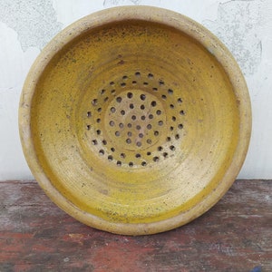 May include: A yellow ceramic colander with a circular shape and a pattern of holes. The colander is sitting on a wooden surface.