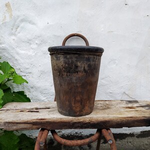 Puede incluir: Un cubo de madera marrón rústico con un asa de metal se sienta sobre una mesa de madera desgastada. El cubo tiene un interior marrón oscuro, casi negro.