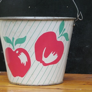 Puede incluir: Un cubo de metal blanco con un diseño de manzana verde y roja. El cubo tiene un asa y está rayado con líneas verdes.