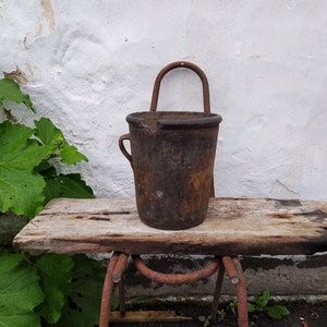May include: A rusty metal bucket with a handle sits on a weathered wooden table. The bucket has a chipped edge and is slightly dented. The table is supported by a rusty metal frame.