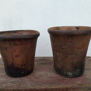 May include: Two terracotta flower pots, one slightly larger than the other, both with a worn, rustic brown finish. The pots are sitting on a wooden surface.