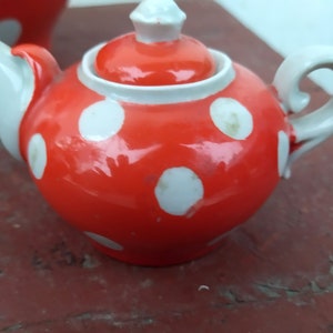 May include: A small, round teapot with a bright red glaze and white polka dots. The spout, handle, and lid are white. The teapot is a charming piece for serving tea.