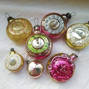 May include: A collection of seven vintage glass Christmas ornaments shaped like clocks. The ornaments are in various colors, including gold, pink, and green. Some of the ornaments have Roman numeral clock faces, while others have Arabic numeral clock faces.