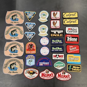May include: A collection of fishing-themed patches and emblems. The patches feature various logos, including "North American Fishing Club," "Culprit," "Abu Garcia," and "Mister Twister." The color palette includes beige, blue, red, and yellow.