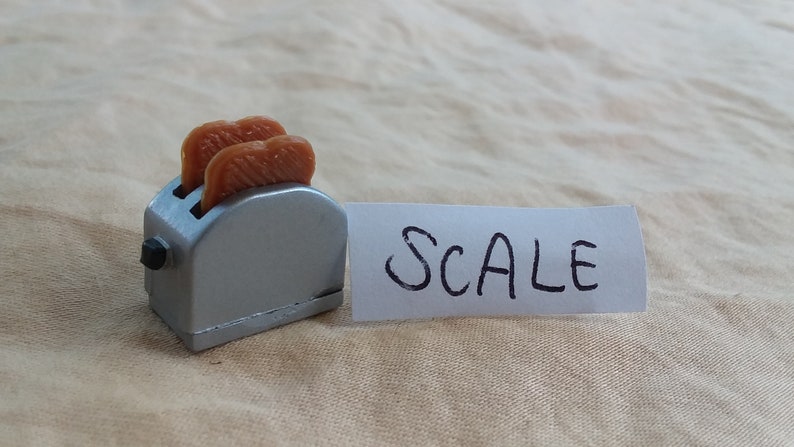 Pu&ograve; includere: Un tostapane argentato in miniatura con due fette di pane all'interno. La parola "SCALE" &egrave; scritta su un pezzo di carta bianco accanto al tostapane.