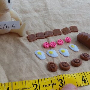 Pu&ograve; includere: Un set di cibo giocattolo in miniatura con una variet&agrave; di articoli, tra cui pane, toast, ciambelle, uova e pasticcini. Il set &egrave; esposto su una superficie di tessuto marrone con un metro a nastro per la scala. Il testo "SCALE" &egrave; scritto su un pezzo di carta bianca.