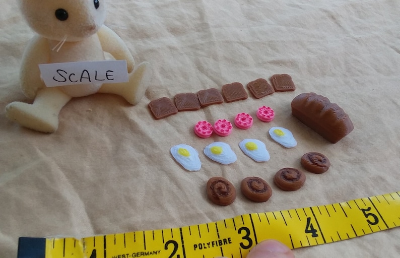 Pu&ograve; includere: Un set di cibo giocattolo in miniatura con una variet&agrave; di articoli, tra cui pane, toast, ciambelle, uova e pasticcini. Il set &egrave; esposto su una superficie di tessuto marrone con un metro a nastro per la scala. Il testo "SCALE" &egrave; scritto su un pezzo di carta bianca.