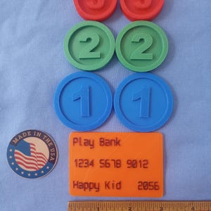 May include: A collection of colorful, 3D-printed play money and accessories. Includes red number 3 coins, green number 2 coins, and blue number 1 coins. Also, an orange "Play Bank" credit card and a wooden ruler.