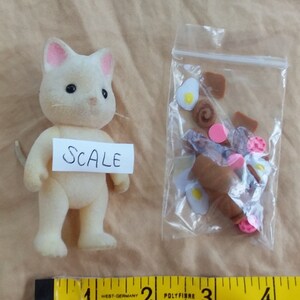 Pu&ograve; includere: Una statuina di gatto bianco con orecchie rosa, in piedi su un tavolo accanto a un sacchetto di plastica trasparente contenente piccoli oggetti alimentari marroni e rosa. Il sacchetto &egrave; etichettato "SCALE". Un metro a nastro giallo &egrave; in primo piano.