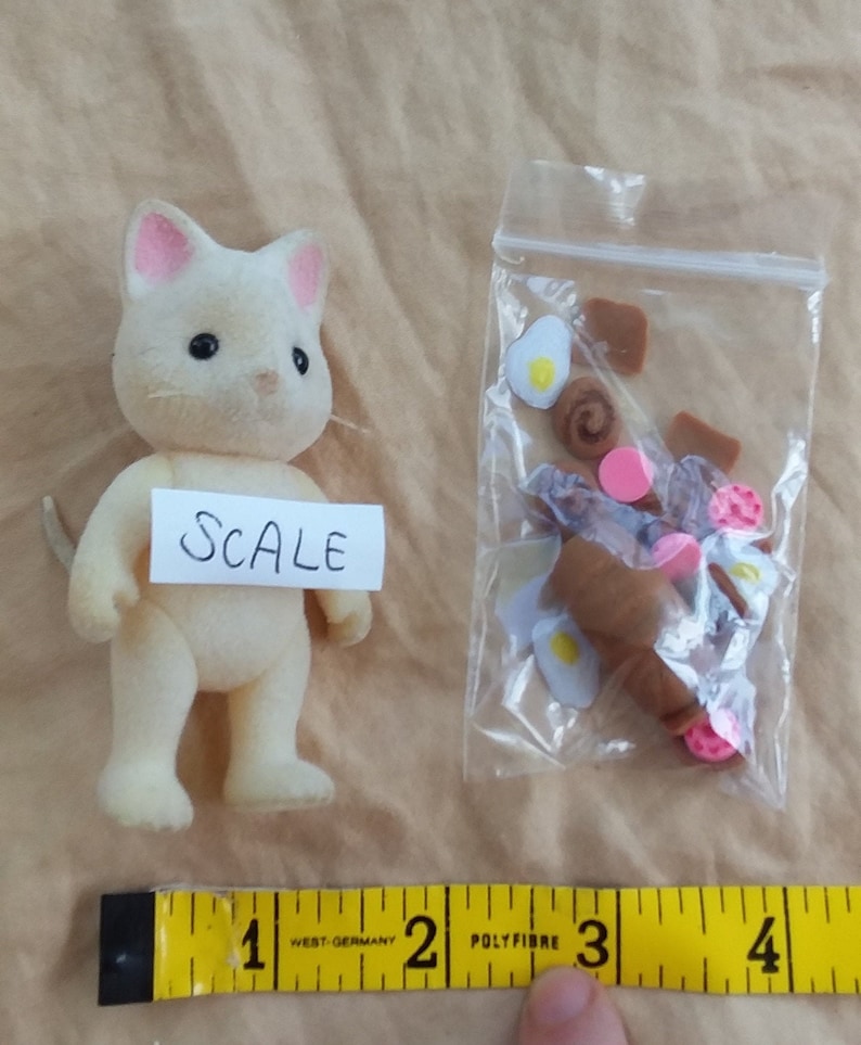 Pu&ograve; includere: Una statuina di gatto bianco con orecchie rosa, in piedi su un tavolo accanto a un sacchetto di plastica trasparente contenente piccoli oggetti alimentari marroni e rosa. Il sacchetto &egrave; etichettato "SCALE". Un metro a nastro giallo &egrave; in primo piano.