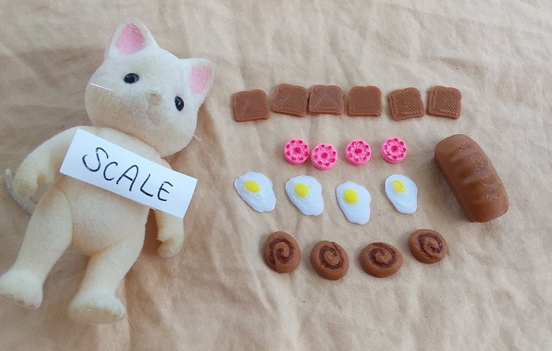 Pu&ograve; includere: Un gatto di peluche bianco con orecchie rosa, che tiene un biglietto bianco che dice "SCALE". L'immagine mostra anche una variet&agrave; di cibi in miniatura, tra cui pane, toast, uova, ciambelle e pasticcini.