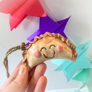 May include: A ceramic dumpling ornament with a smiling face and heart-shaped cheeks. The dumpling is tan with a crimped edge and a jute string for hanging. Colourful paper stars are in the background.