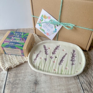May include: A gift set comprising a white ceramic soap dish with lavender design, a lavender soap bar, and a natural soap saver. The set is tied with green twine and includes a cardboard box and a tag that reads "Soap Dish Gift Set".