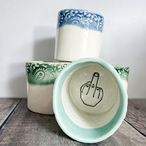 May include: A collection of ceramic cups with a smooth white base and a textured, embossed rim in blue or green. One cup is positioned in the foreground, revealing a black line drawing of a hand making a middle finger gesture on the interior base.